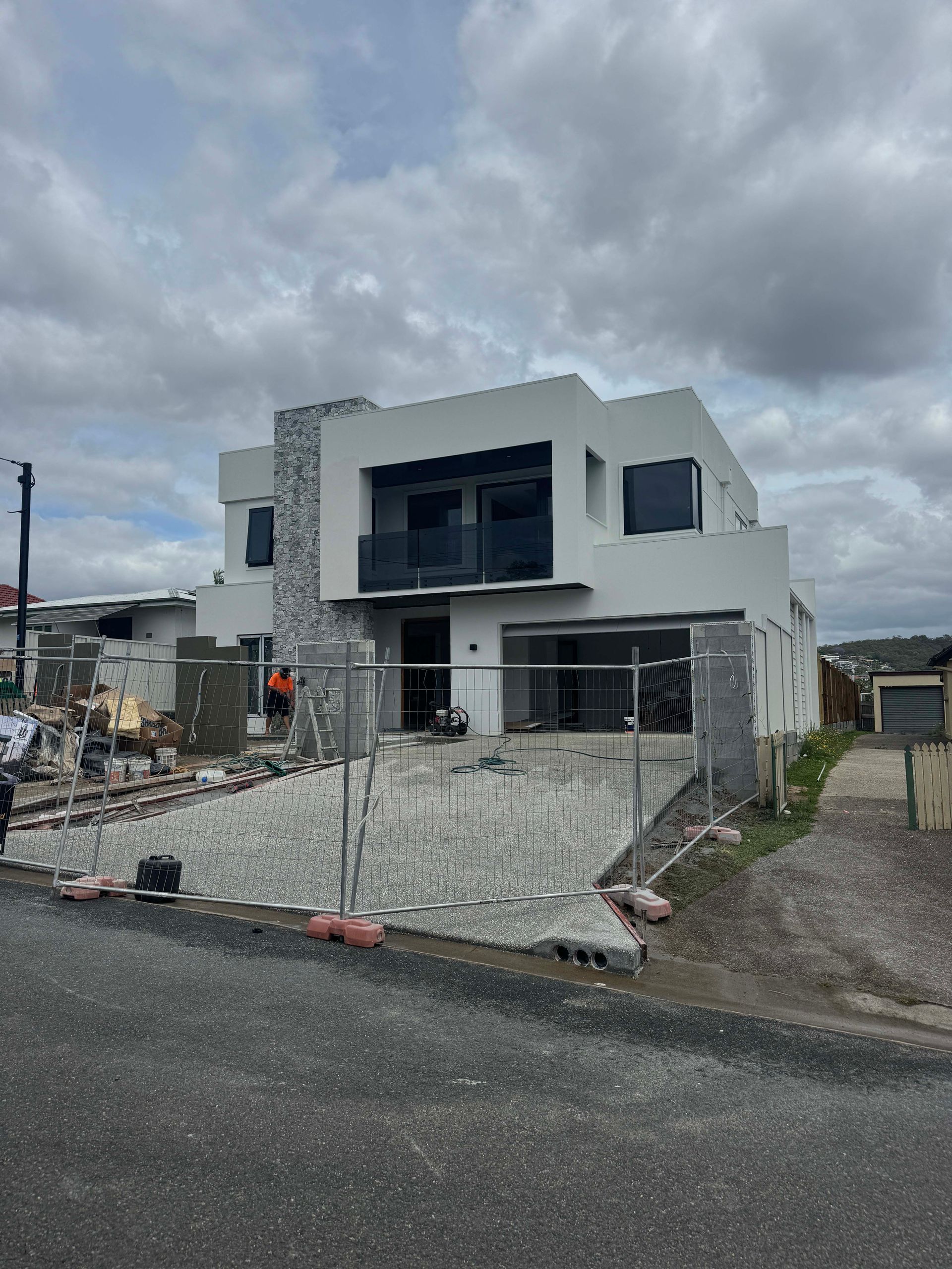Two-story modern home under construction with light gray facade, stone accents, and a balcony, on a gravel lot.