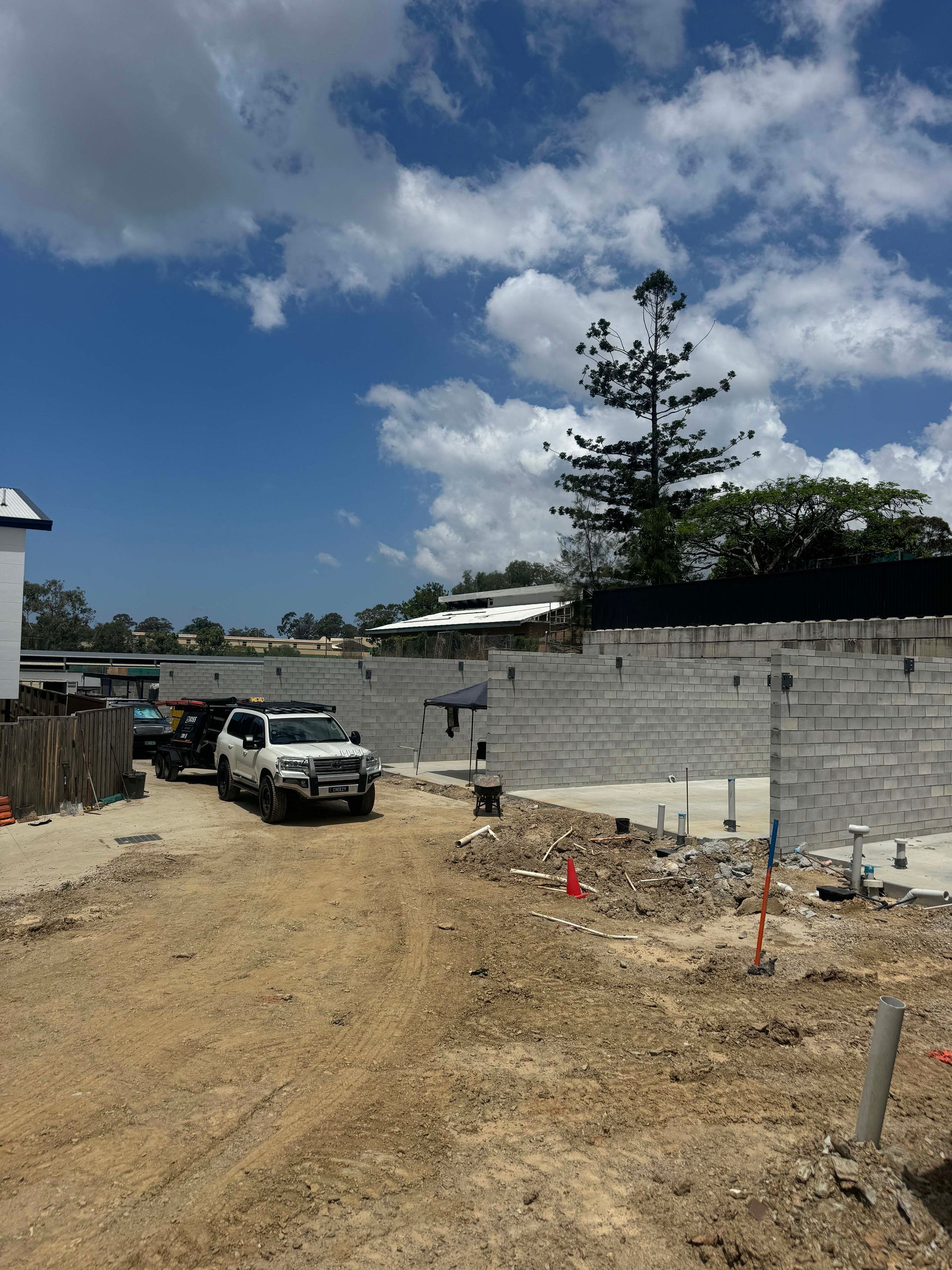 Construction site with a white SUV pulling a trailer. Gray concrete block wall, blue sky with white clouds.
