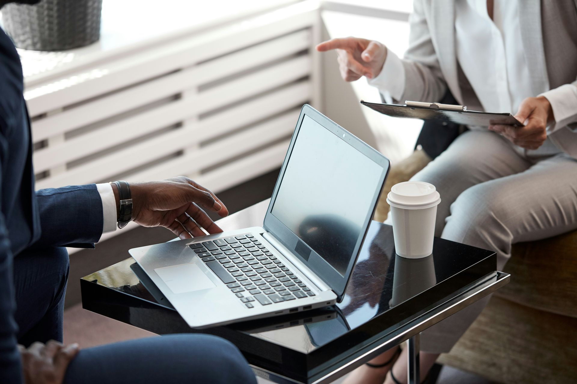 Two people in business attire at a table with a laptop, pointing at a clipboard, coffee cup.