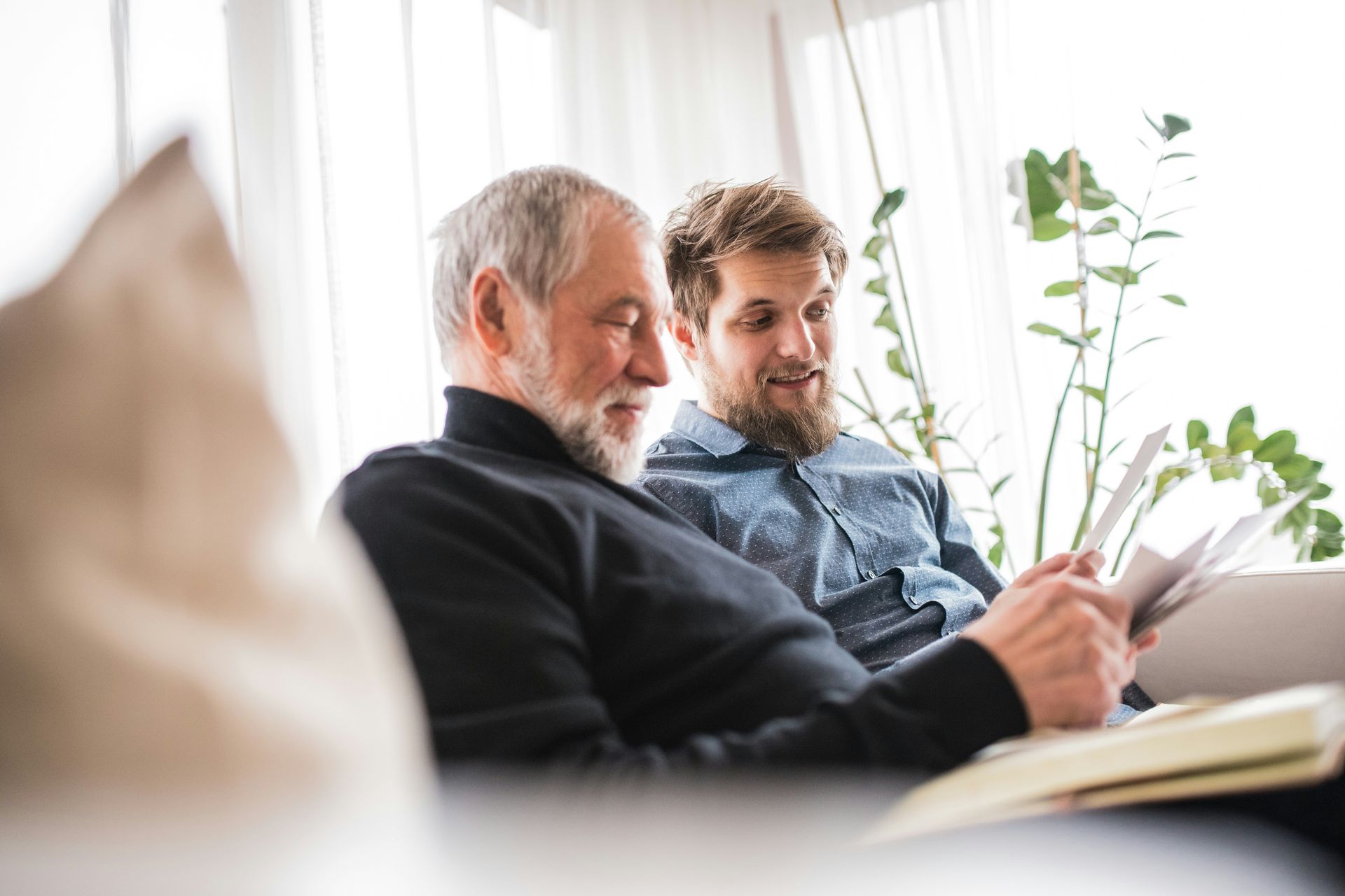 An older man and a younger man sit on a couch, looking at a book together in a bright room.