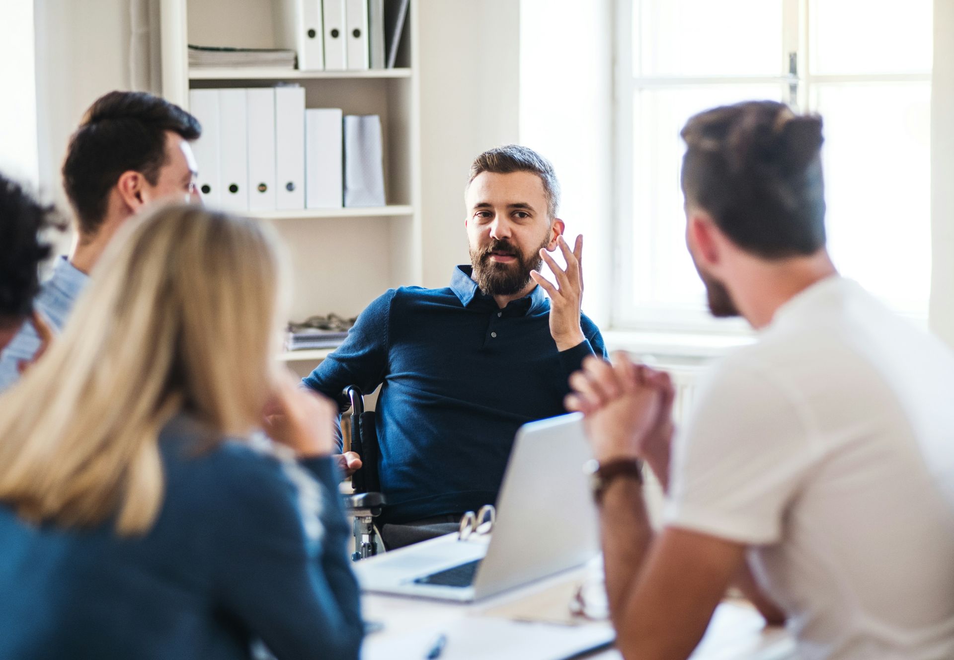 People in a meeting around a table with a laptop, discussing. Man in blue shirt gestures while talking.
