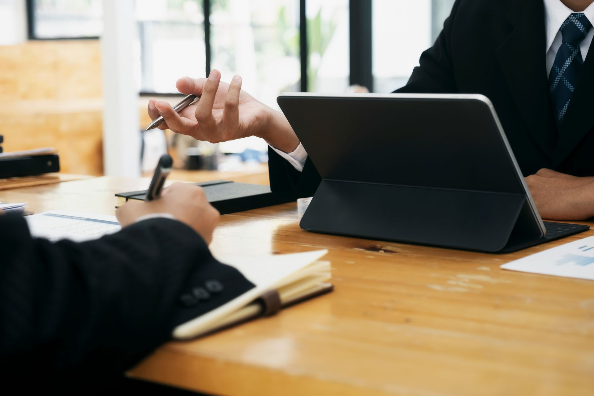 Two people in suits at a wooden table; one points a pen, the other takes notes next to a tablet.