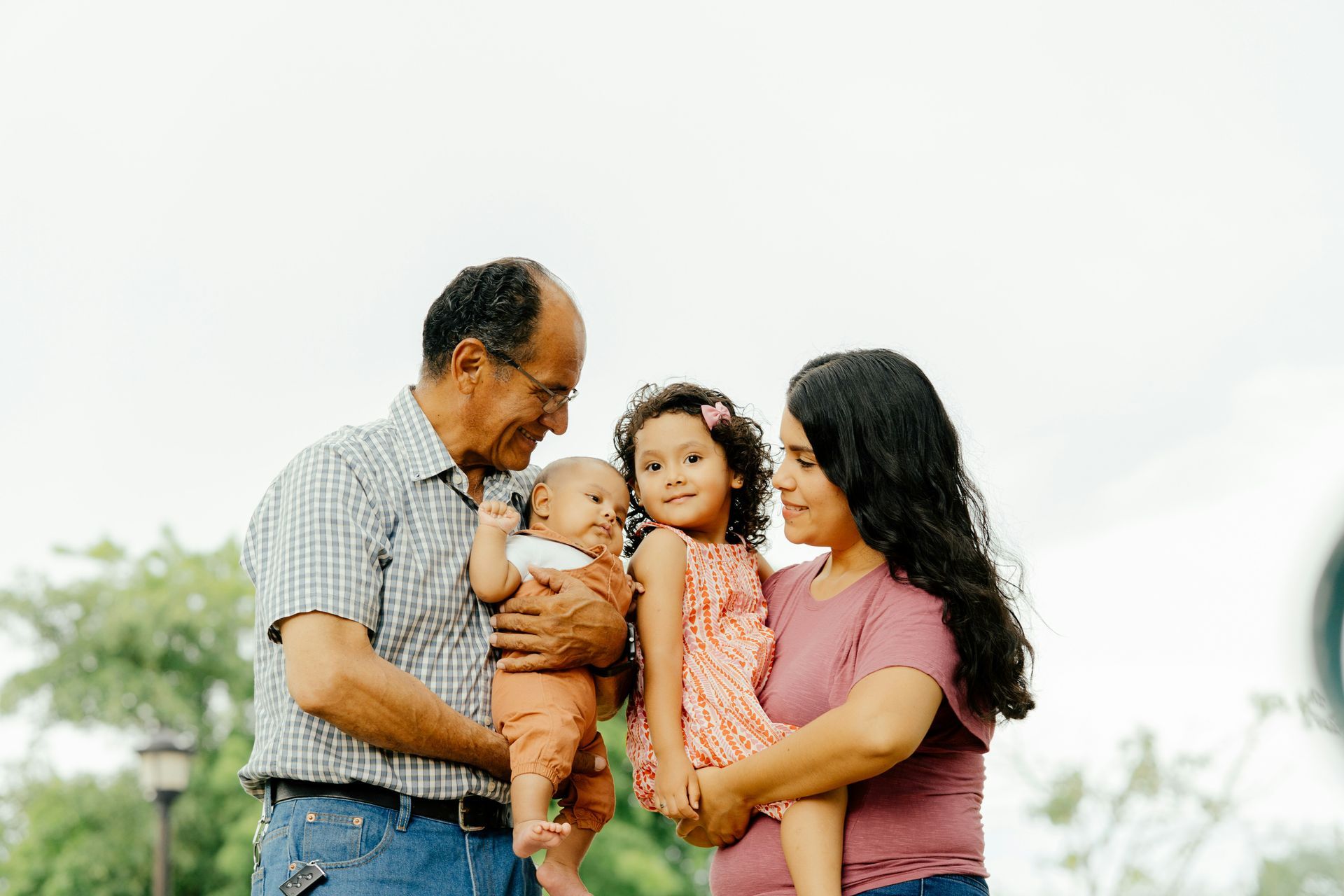 Family outdoors: older adult holding a baby, woman holding a young child, all smiling. Cloudy sky.