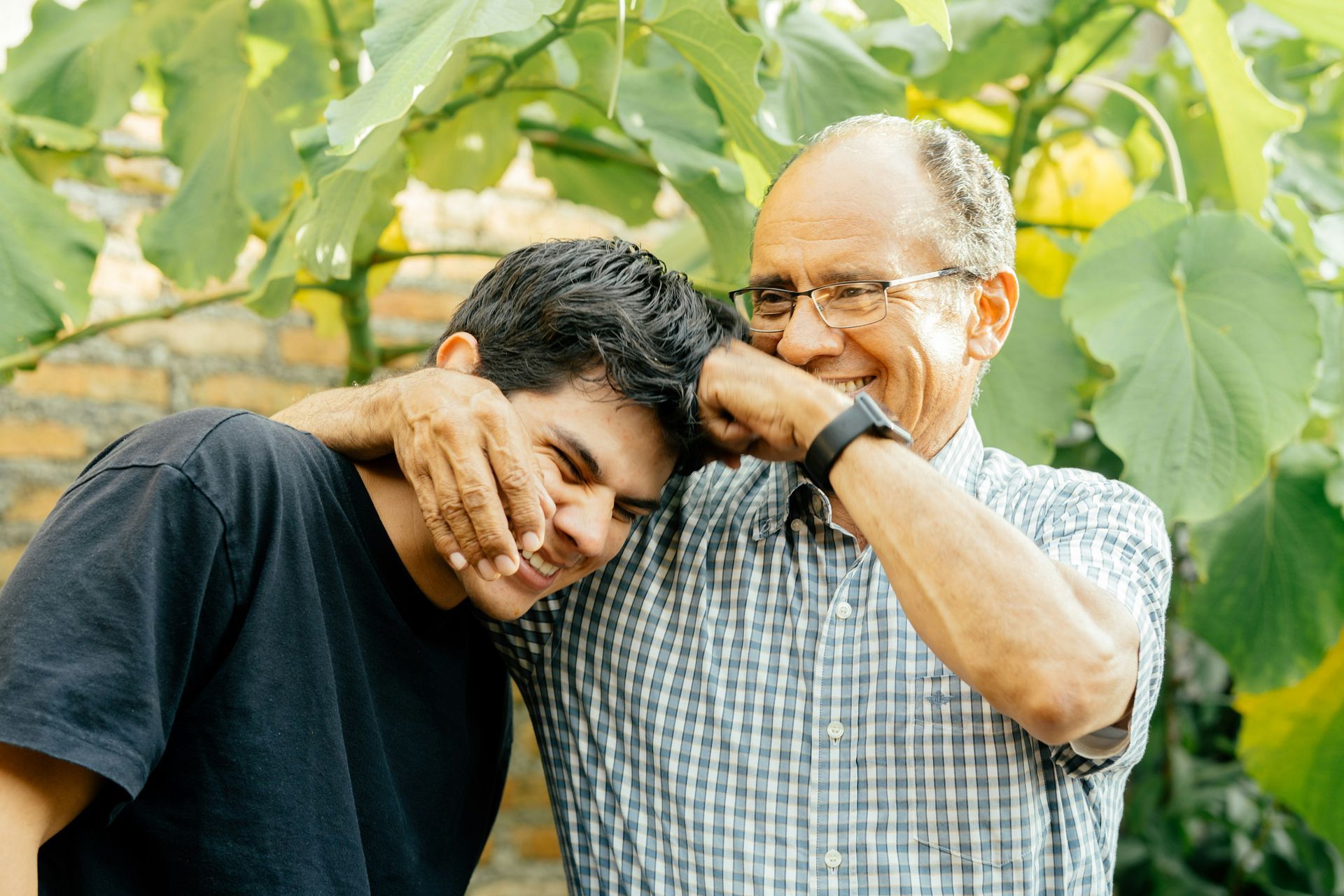 Man with arm around another man, both smiling near greenery.