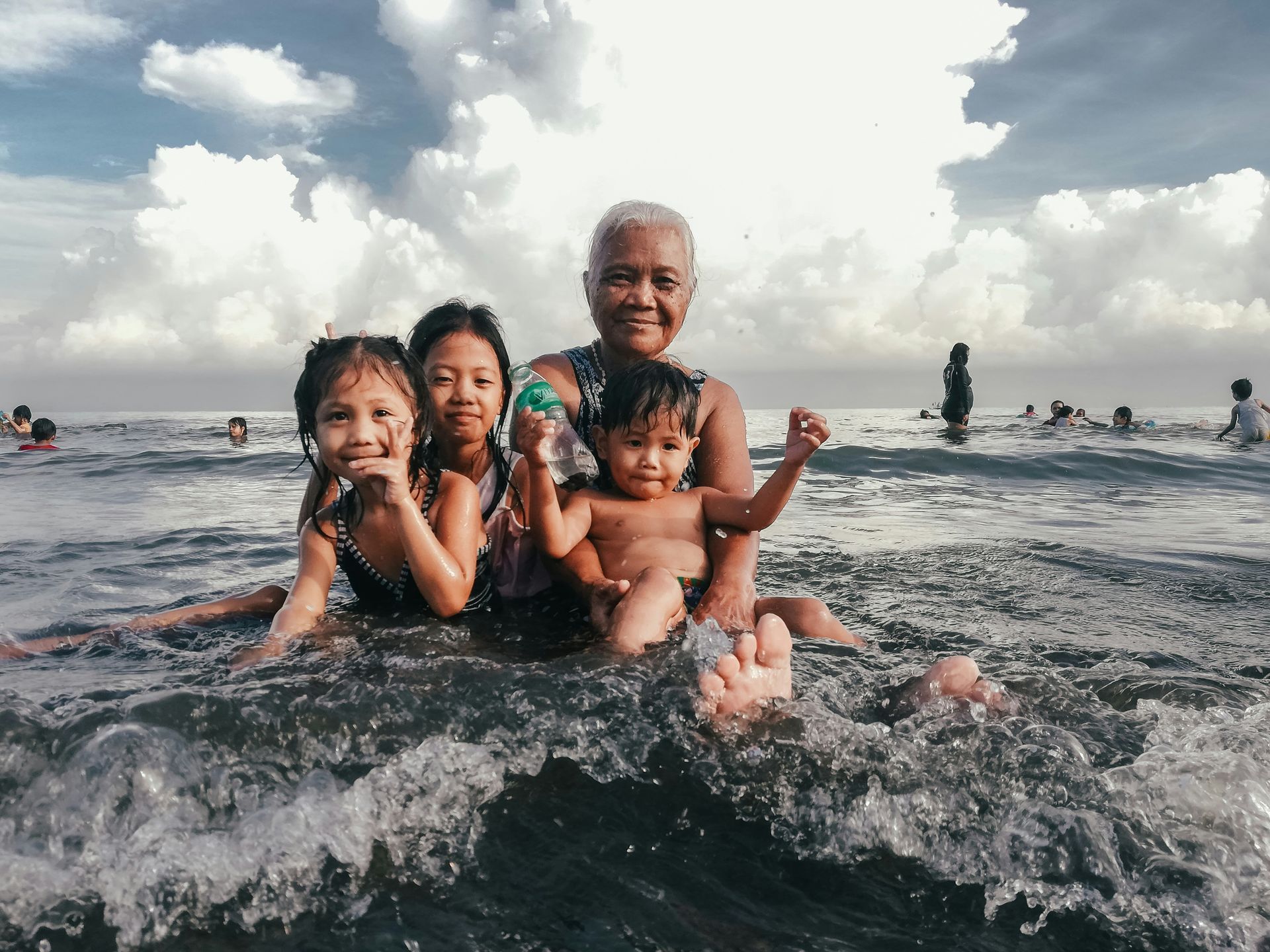 A woman and three children in ocean water, waves splashing. Cloudy sky overhead.
