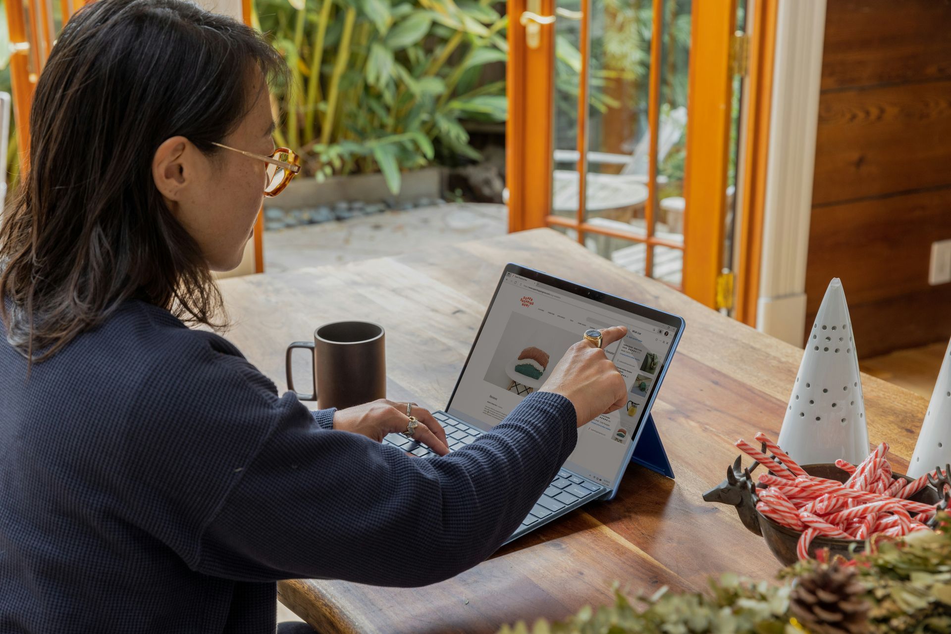 Woman using a tablet on a wooden table, touching the screen. Coffee mug and holiday decor nearby.