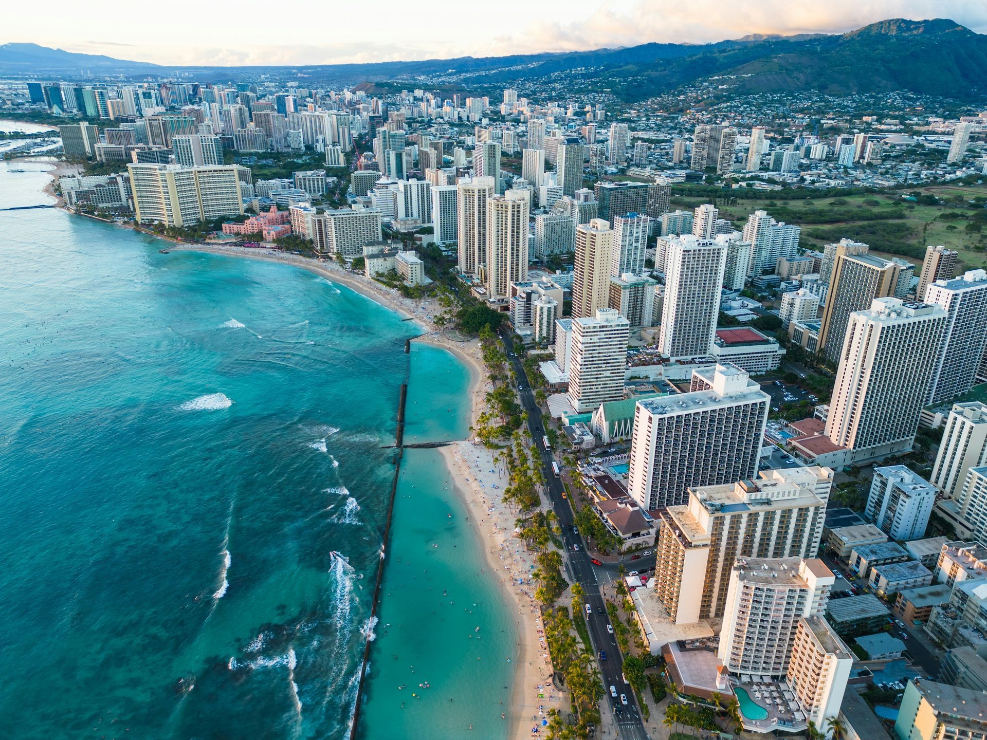 Waikiki Beach, Honolulu. Aerial view of city skyline, turquoise ocean, white sand beach, and green mountains under blue sky.