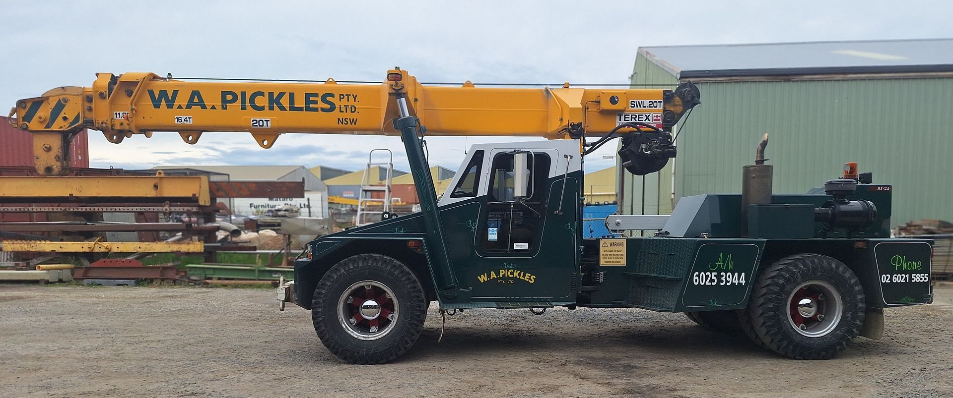 Yellow crane on green truck in a gravel lot, with another truck in the background
