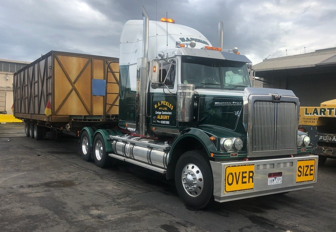 Dark green semi-truck hauling a covered trailer in a wet industrial lot, with yellow “OVER” signs on the bumper