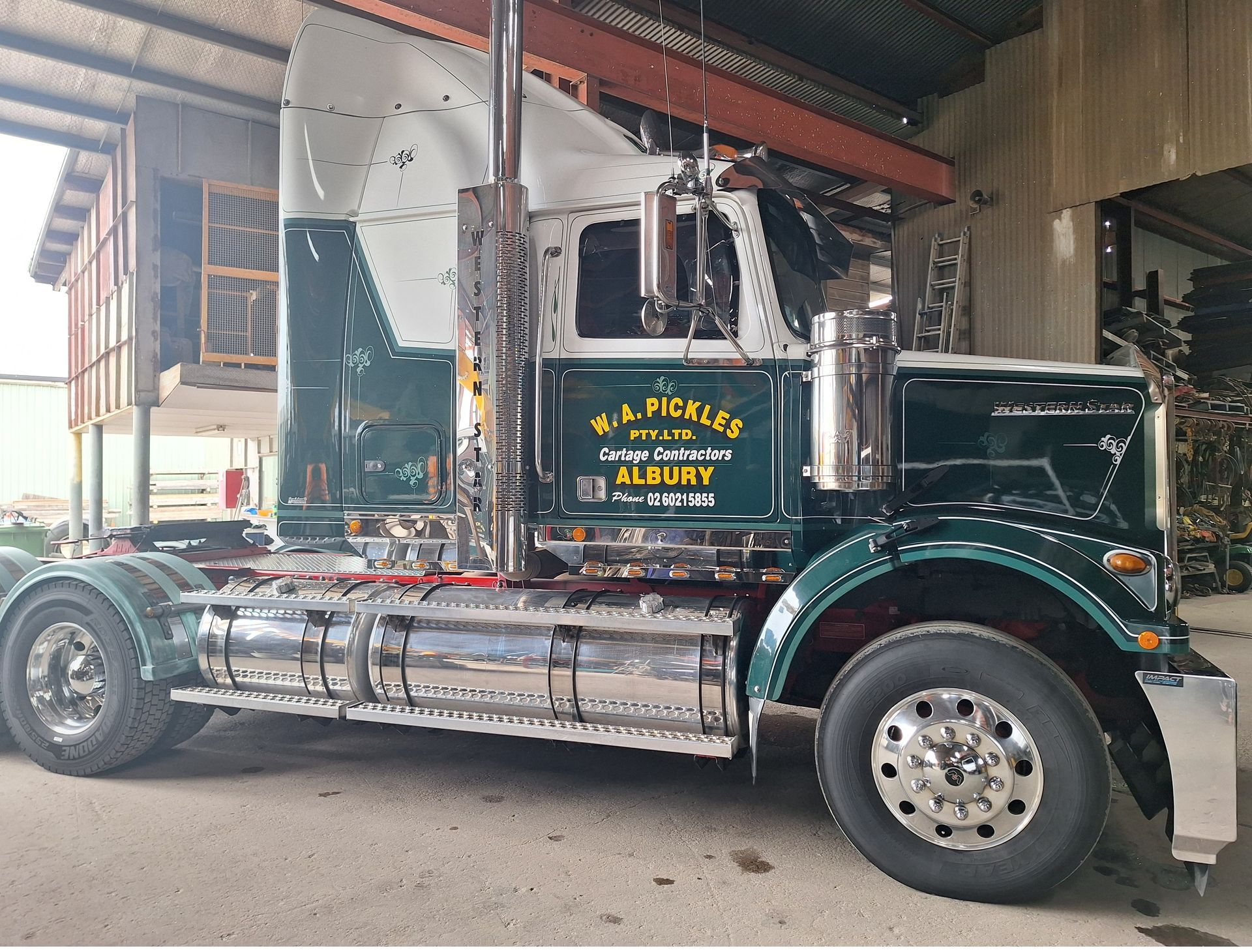 Dark green Mack truck with chrome tank trailer parked under a warehouse roof, side view