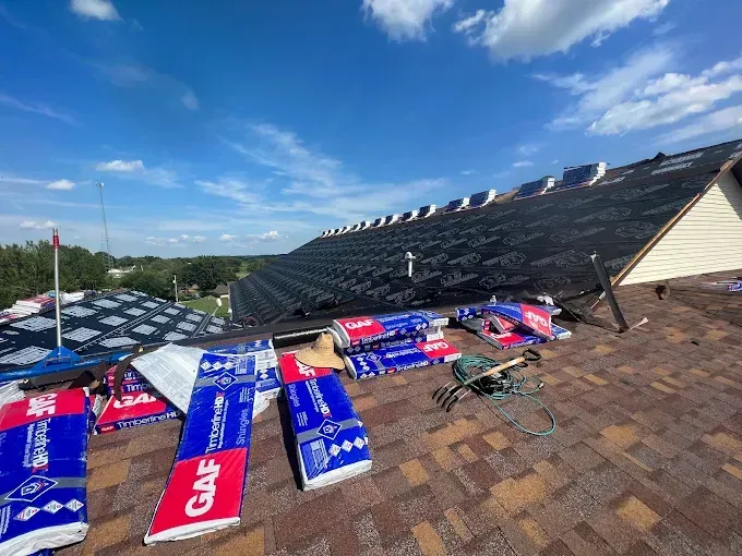 Roof partially covered with asphalt shingles. GAF shingles and tools are scattered on the roof. Bright blue sky.
