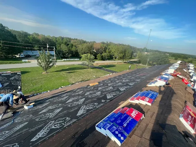 Roofers working on a residential roof, asphalt shingles visible. Blue sky, green grass, and trees in the background.