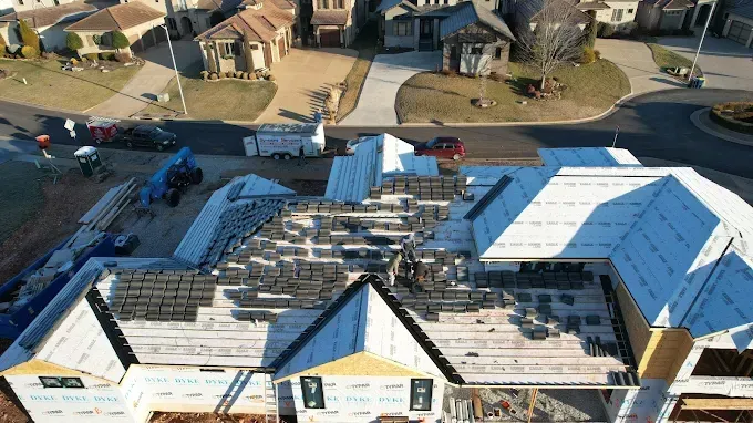 Rooftop construction: workers installing dark gray tiles on a large house. Blue tarp, wooden beams, sunny day.
