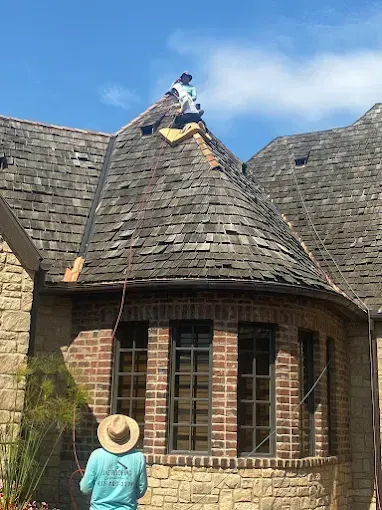 Two roofers repairing a weathered, shingle roof on a brick and stone building under a blue sky.