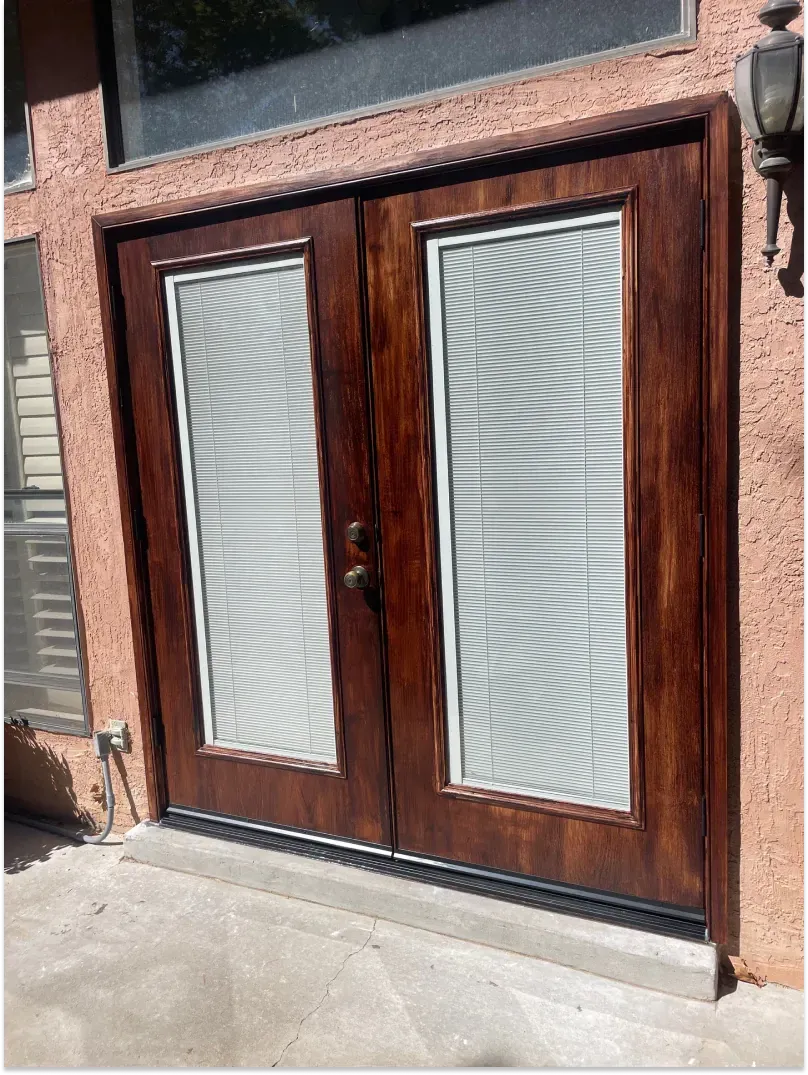 Double wooden doors with glass panes and blinds, set in a reddish-brown stucco exterior.