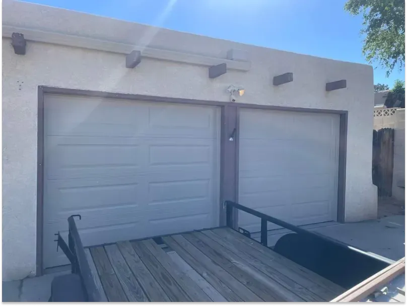 Two-car garage with white doors, light-colored stucco exterior, and a trailer parked in front.