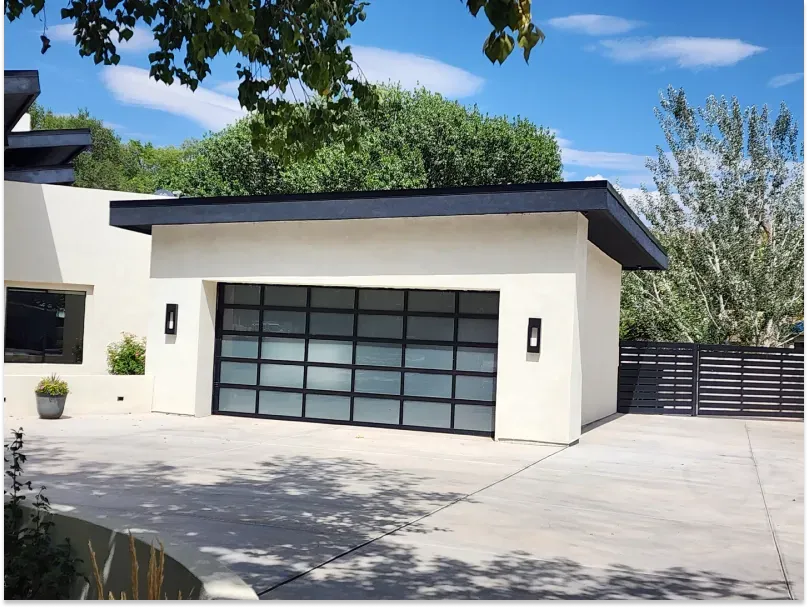 Modern garage with frosted glass door, black trim, and off-white exterior. Concrete driveway, lush greenery.
