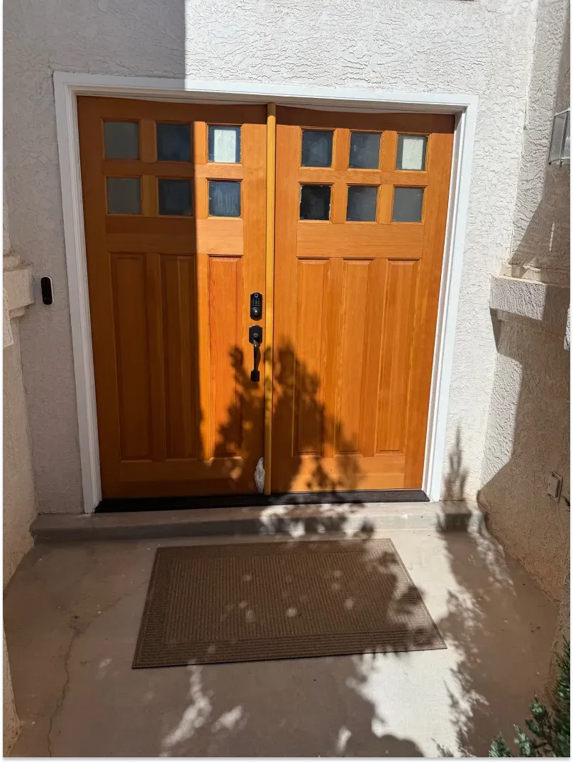 Wooden double doors with square glass panes above, on a concrete porch, with a welcome mat; shadow of tree.