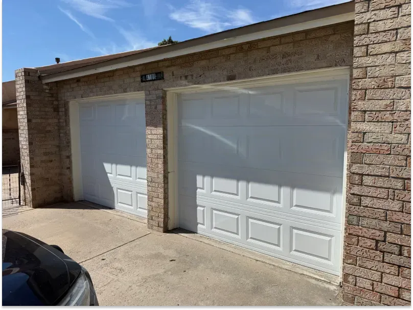 Two white garage doors in brick building under a blue sky. A car is parked on the left.