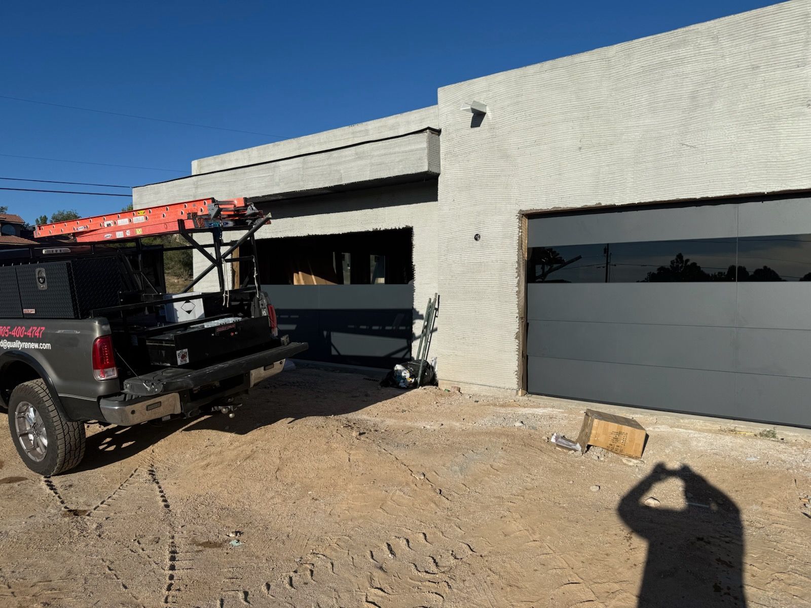 A gray truck parked near a modern gray garage. A person's shadow is visible, holding a phone.