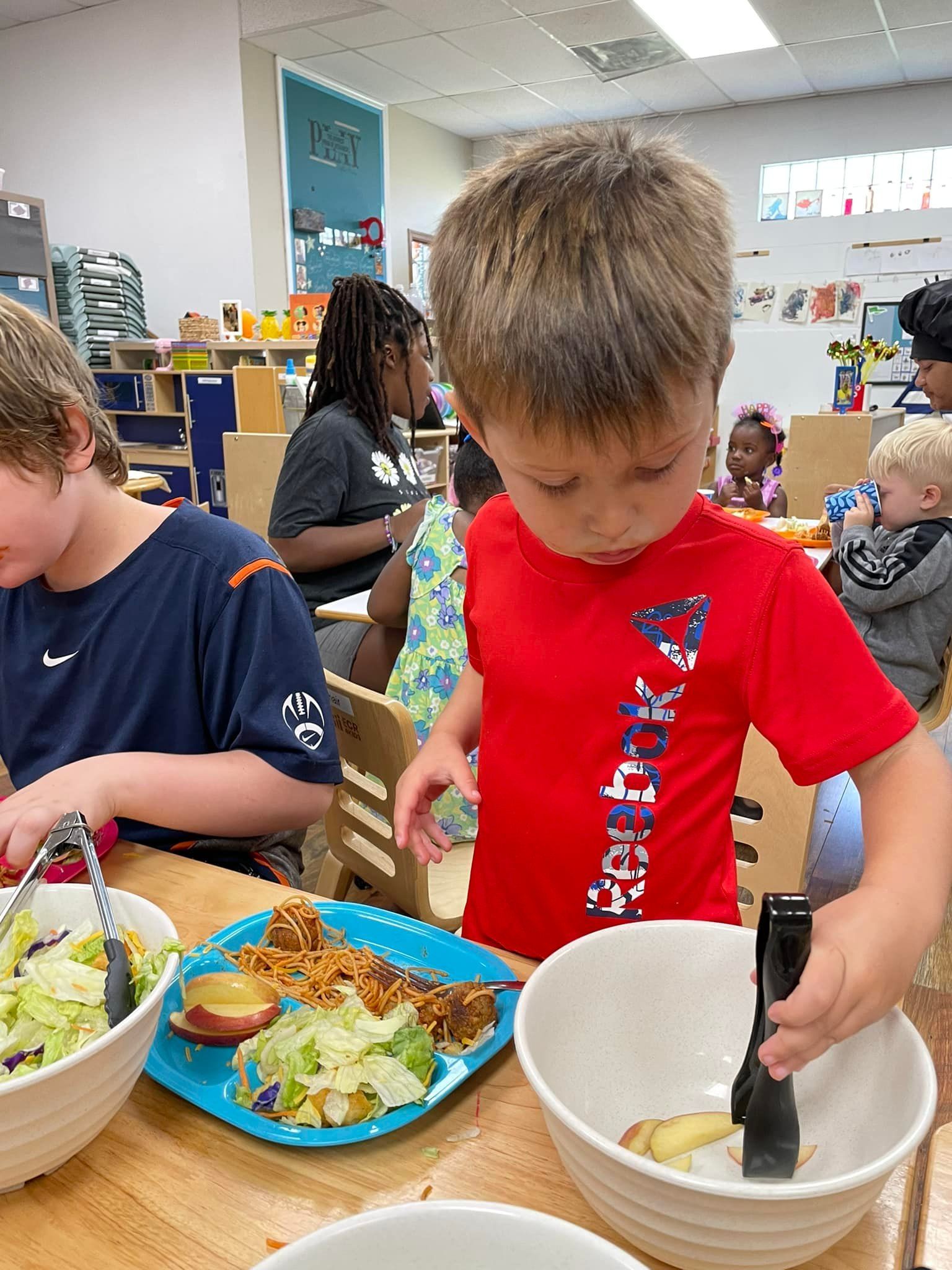 Young boy in red shirt serving food at a table. Other children eat lunch in the background.