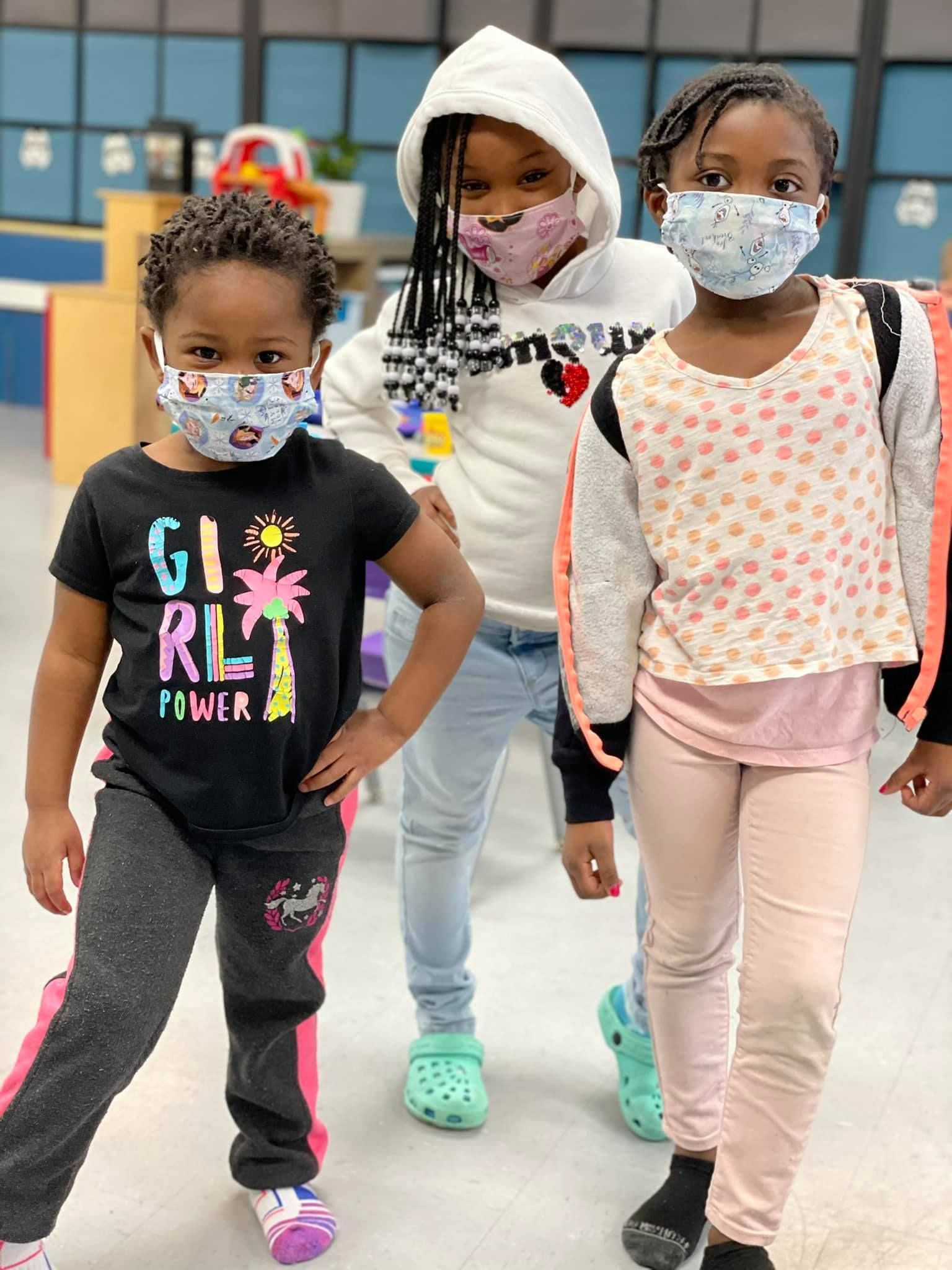 Three young Black girls wearing masks, casual clothes, standing indoors, posing.