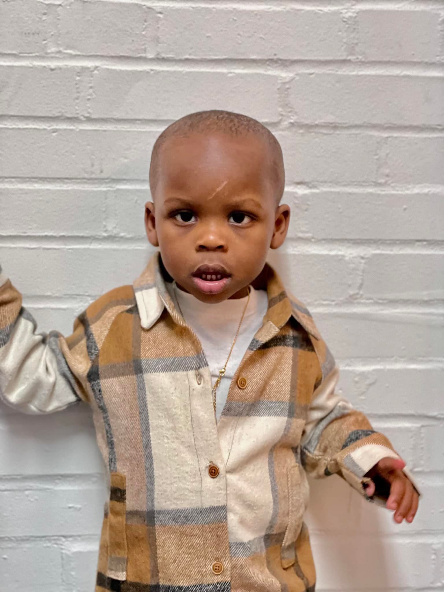 Young child with a shaved head, wearing a plaid shirt and white shirt, standing against a white brick wall.