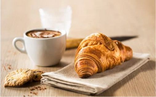 Croissant on a napkin, coffee in a white mug, cookie, and a glass of water on a wooden table.
