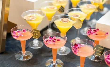 Cocktails in stemmed glasses, some pink with flower petals, others yellow with citrus garnish, on a table.