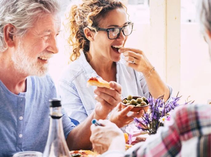 People at a table, smiling and eating food. One holds olives, another bread. Indoor setting.