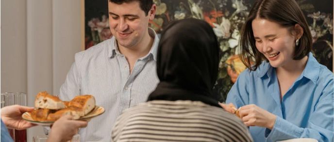 People at a table, smiling. One person offers bread. Another is eating. Blurred floral background.