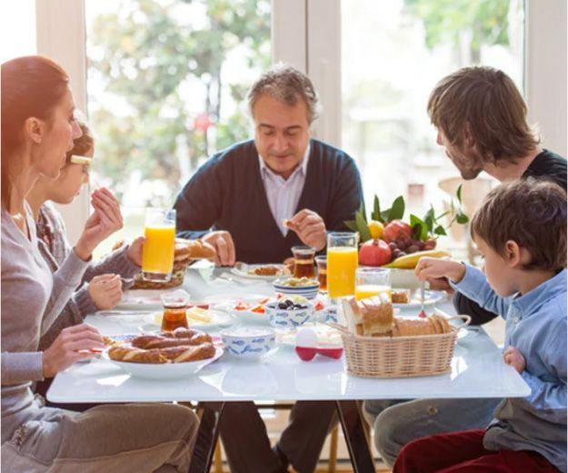 Family eating breakfast at a table near a window; includes juice, bread, and fruit.