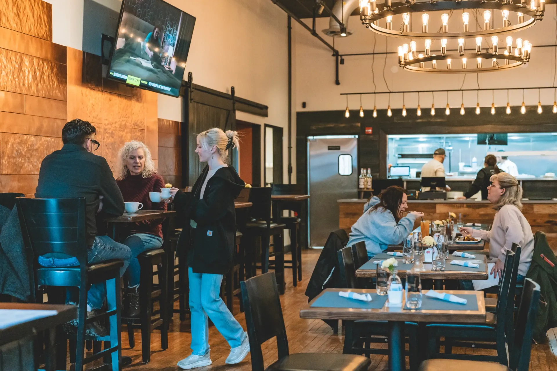 Restaurant interior: patrons seated at tables, bar, kitchen view, neutral tones, warm lighting.