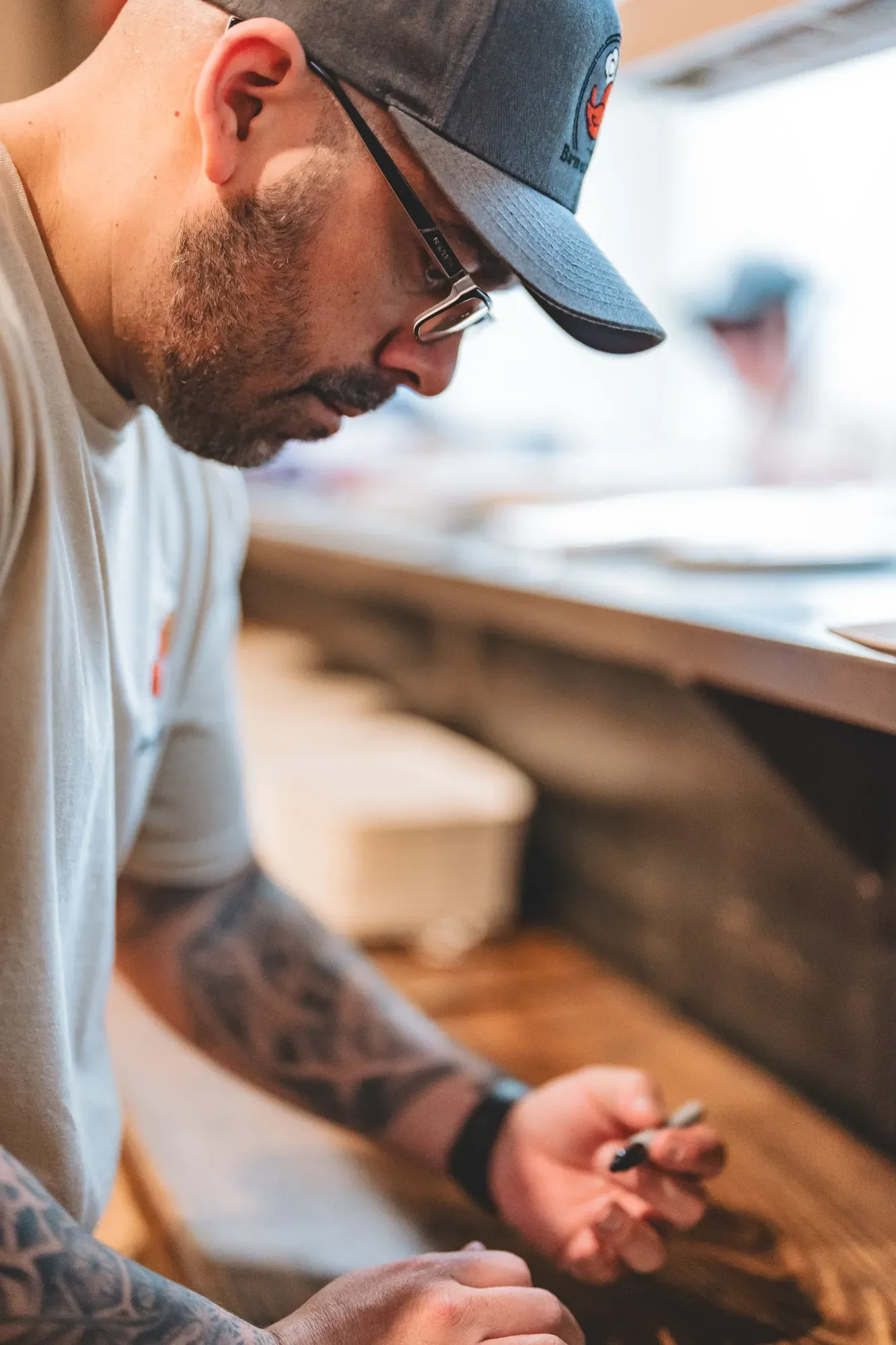 Man with a beard and baseball cap leaning over a counter, holding a pen.