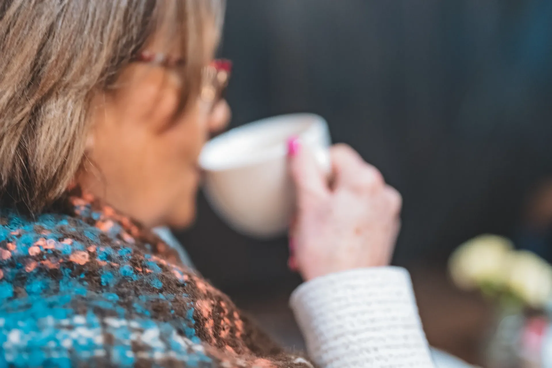 Woman sipping from a white cup. Wearing glasses, blue and brown patterned shawl, and white sweater.