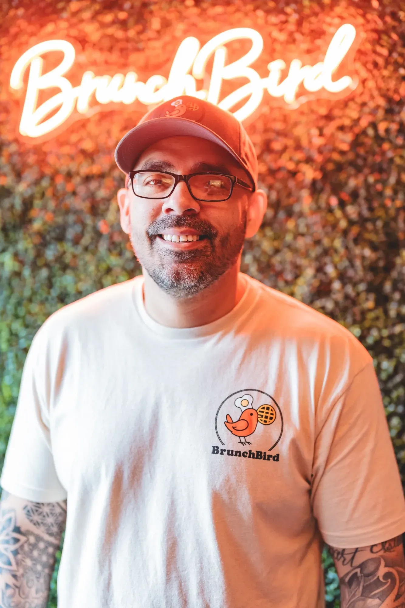 Man wearing a cap and glasses, smiling in front of a neon 