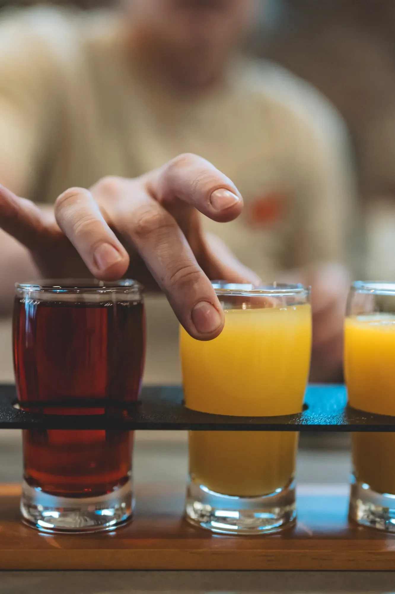 Hand reaching for a shot of yellow liquid; two other shots of different colored liquid on wooden tray.