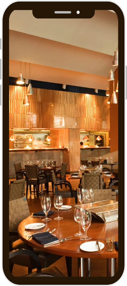 A restaurant interior viewed through a phone screen. A table is set with glassware, napkins, and plates.