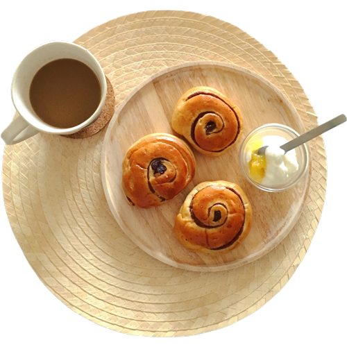 Breakfast: coffee in mug, cinnamon rolls, and yogurt on a wooden plate, on a woven mat.