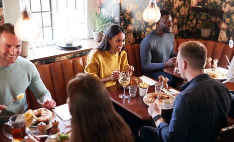 Friends laughing and eating at a restaurant table. Warm lighting, booth seating, food, drinks visible.