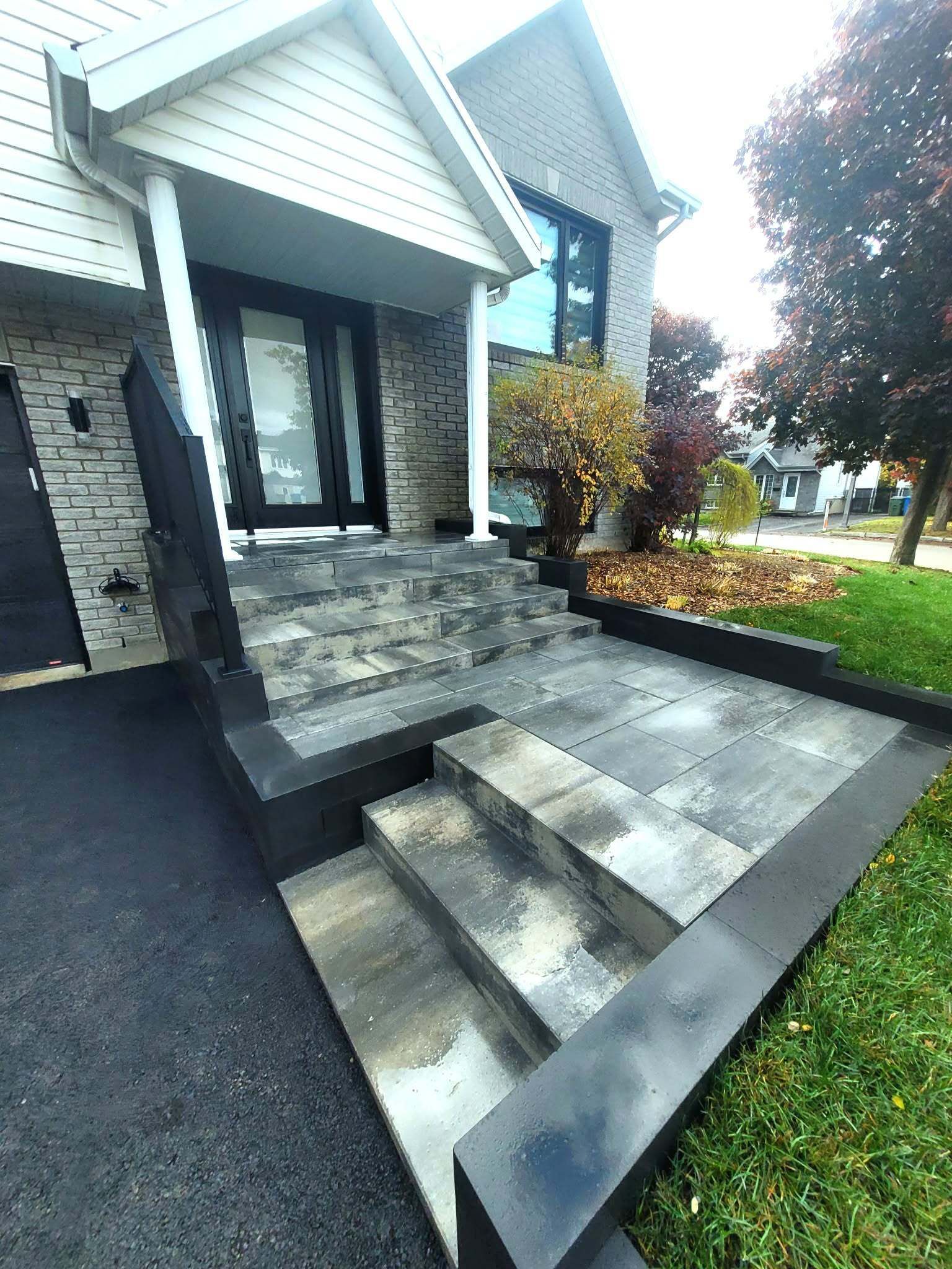 Escaliers et allée en pierre menant à l'entrée de la maison. Bordures noires, allée asphaltée, pelouse verte et arbres.