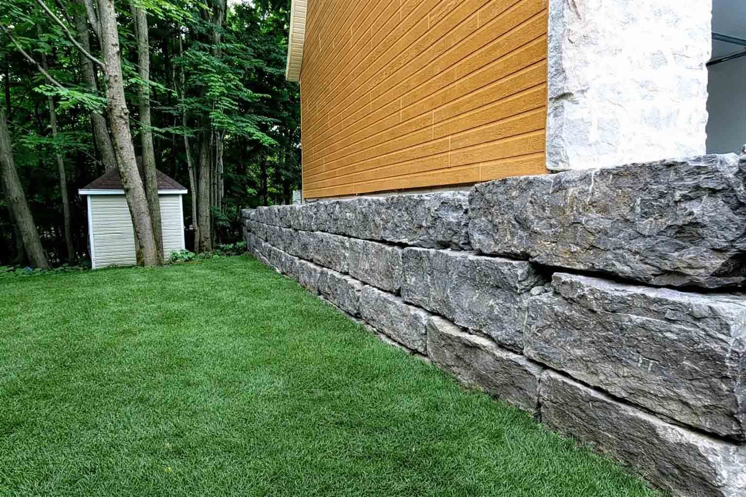 Mur de soutènement en pierre devant un bâtiment au bardage jaune et à la pelouse verte.