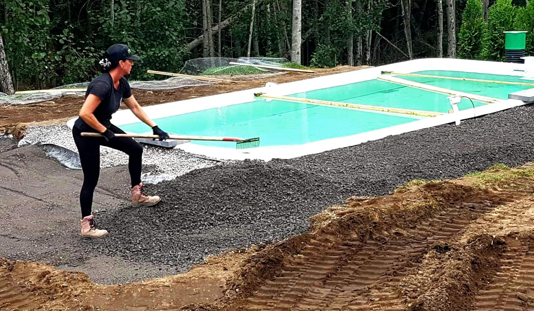 Une femme ratisse le gravier autour d'une piscine rectangulaire en construction dans une zone boisée.