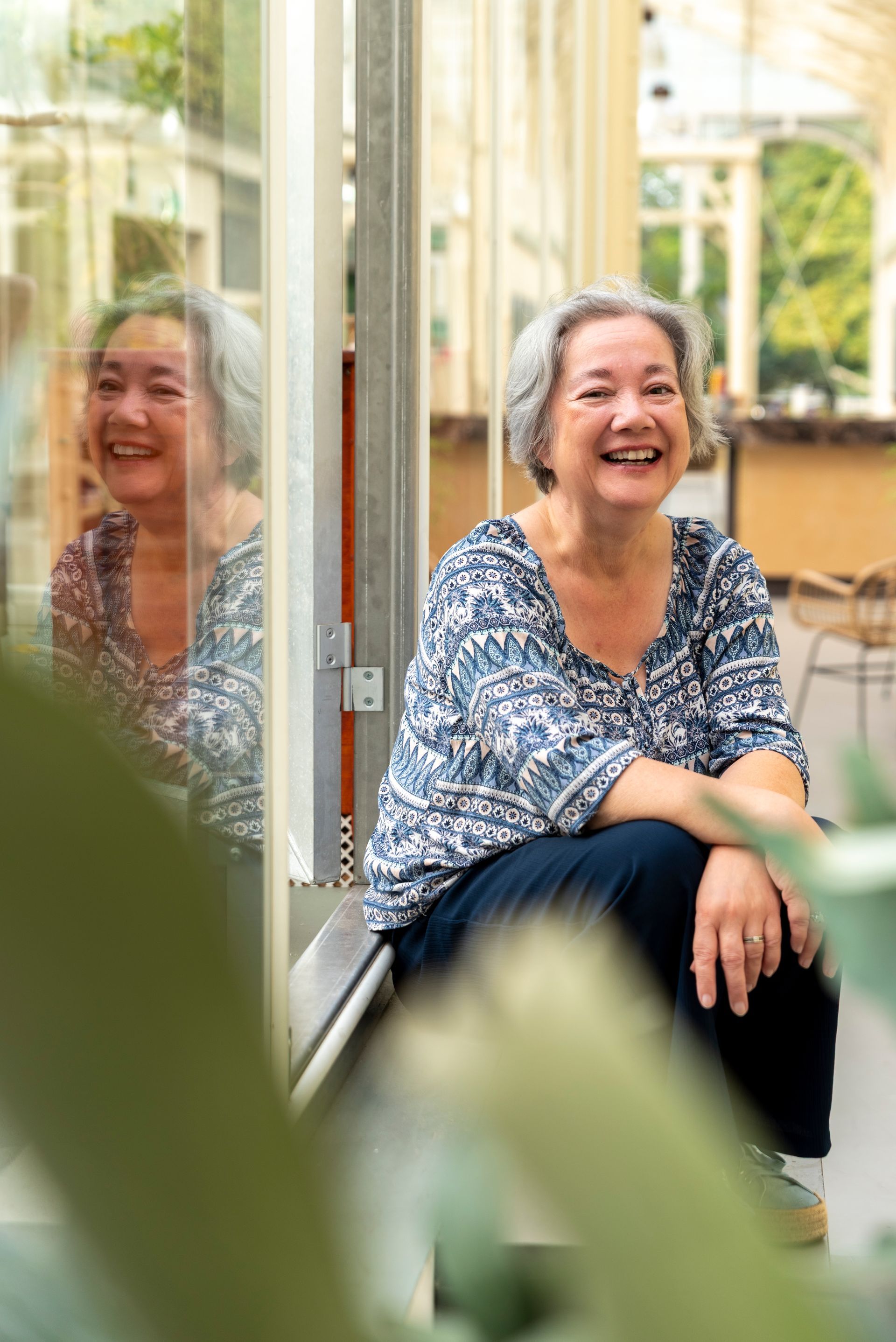 Een vrouw in een donkerblauwe blouse en jeans glimlacht, leunend tegen een vensterbank met planten en een raam op de achtergrond.