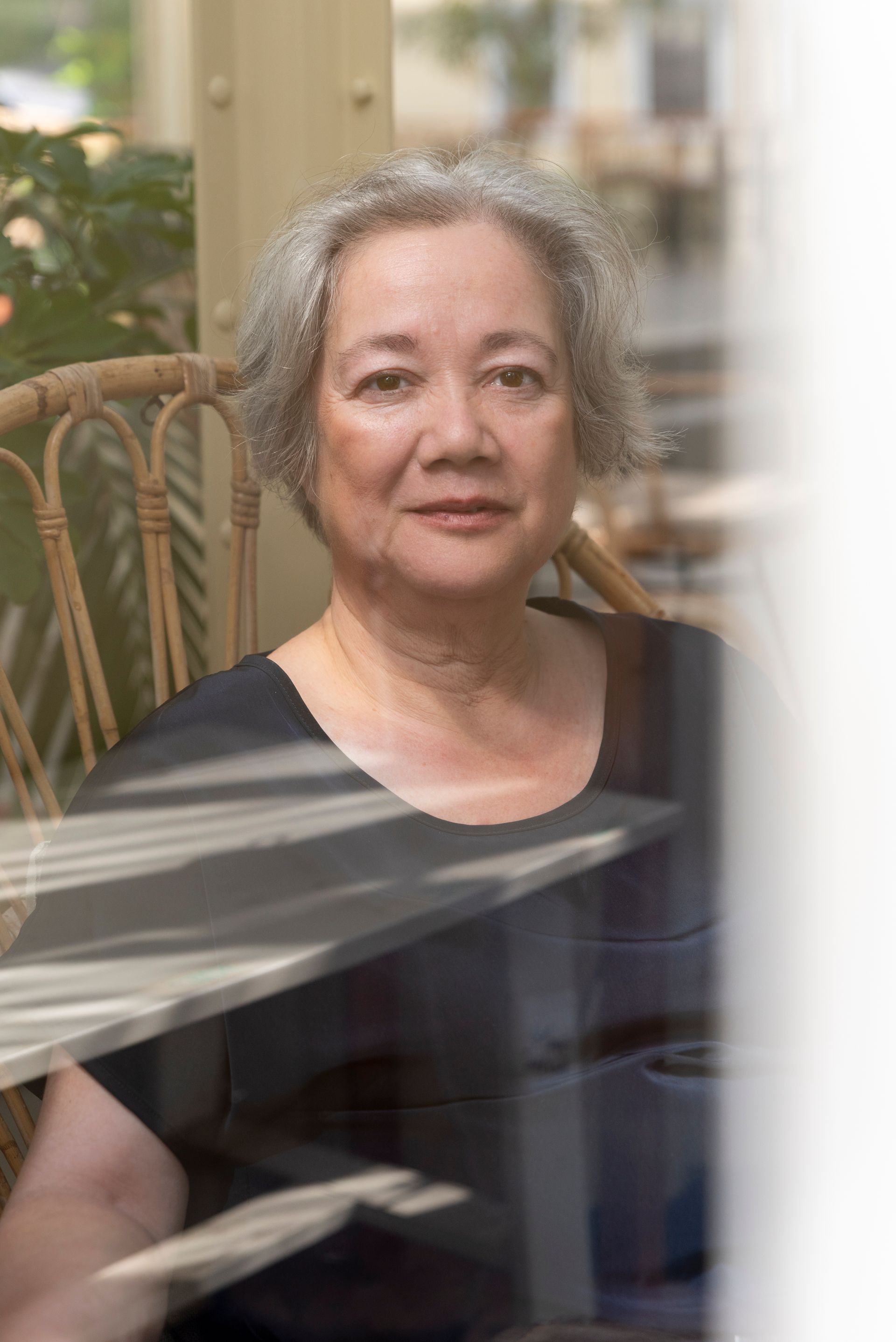 Vrouw met grijs haar zit in een rieten stoel en kijkt voor zich uit; binnen, zonlicht.