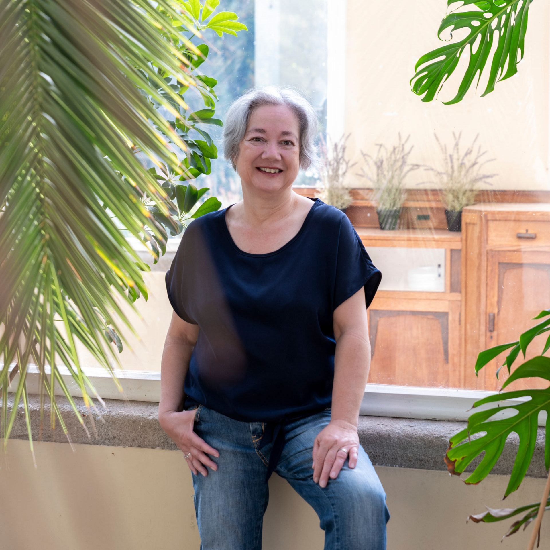 Een vrouw in een donkerblauwe blouse en jeans glimlacht, leunend tegen een vensterbank met planten en een raam op de achtergrond.