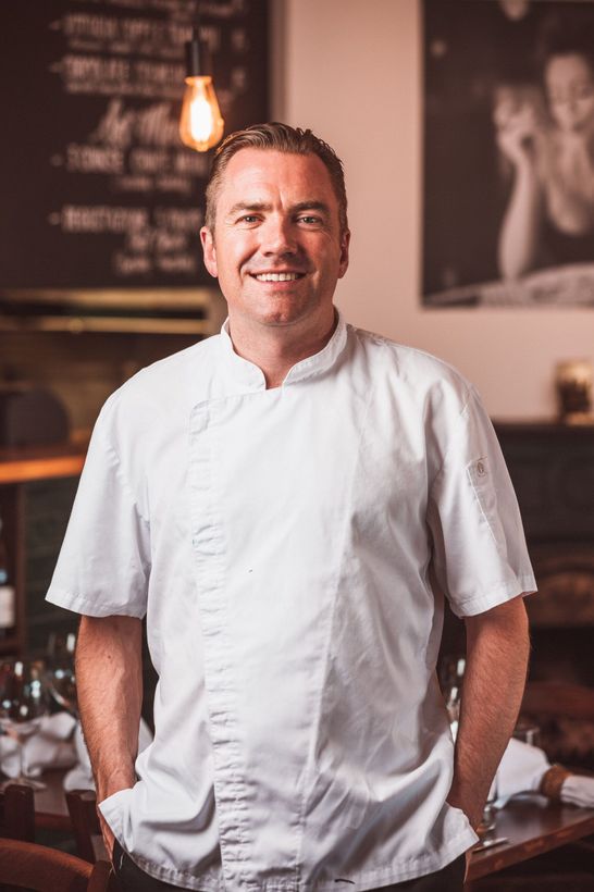 a man in a white chef 's jacket is standing in a restaurant with his hands in his pockets .