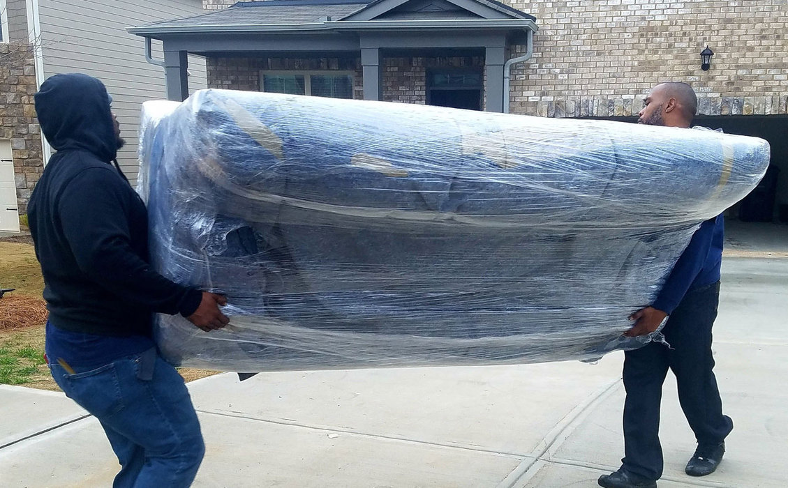 Two people carrying a large, plastic-wrapped couch on a driveway in front of a house.