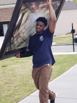 Man carrying a large framed picture outdoors. He wears a navy shirt and brown pants, carrying picture above head.