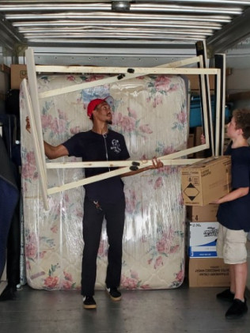 Movers loading furniture into a moving truck; one man holds a bed frame while another carries a box.